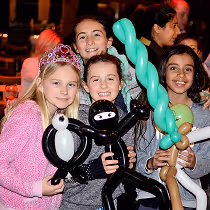 Four children smiling and holding balloon animals and figures at an indoor event, with warm lighting in the background.