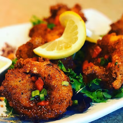 Plate of crispy fried shrimp garnished with chopped green onions and red peppers, served on a bed of leafy greens with a lemon wedge on top.