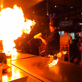 Chef in a restaurant performing a flaming cooking show on a teppanyaki grill, with large flames shooting up as diners watch in the background.