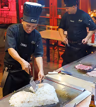 Two chefs wearing black uniforms and hats are cooking on a large flat grill; one chef is using cooking utensils to prepare food while the other looks on.