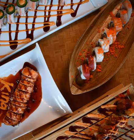 Close-up of chopsticks holding a sushi roll with red tuna, white rice, avocado, and a dark filling, topped with bright orange fish roe, with more sushi and roe blurred in the background.