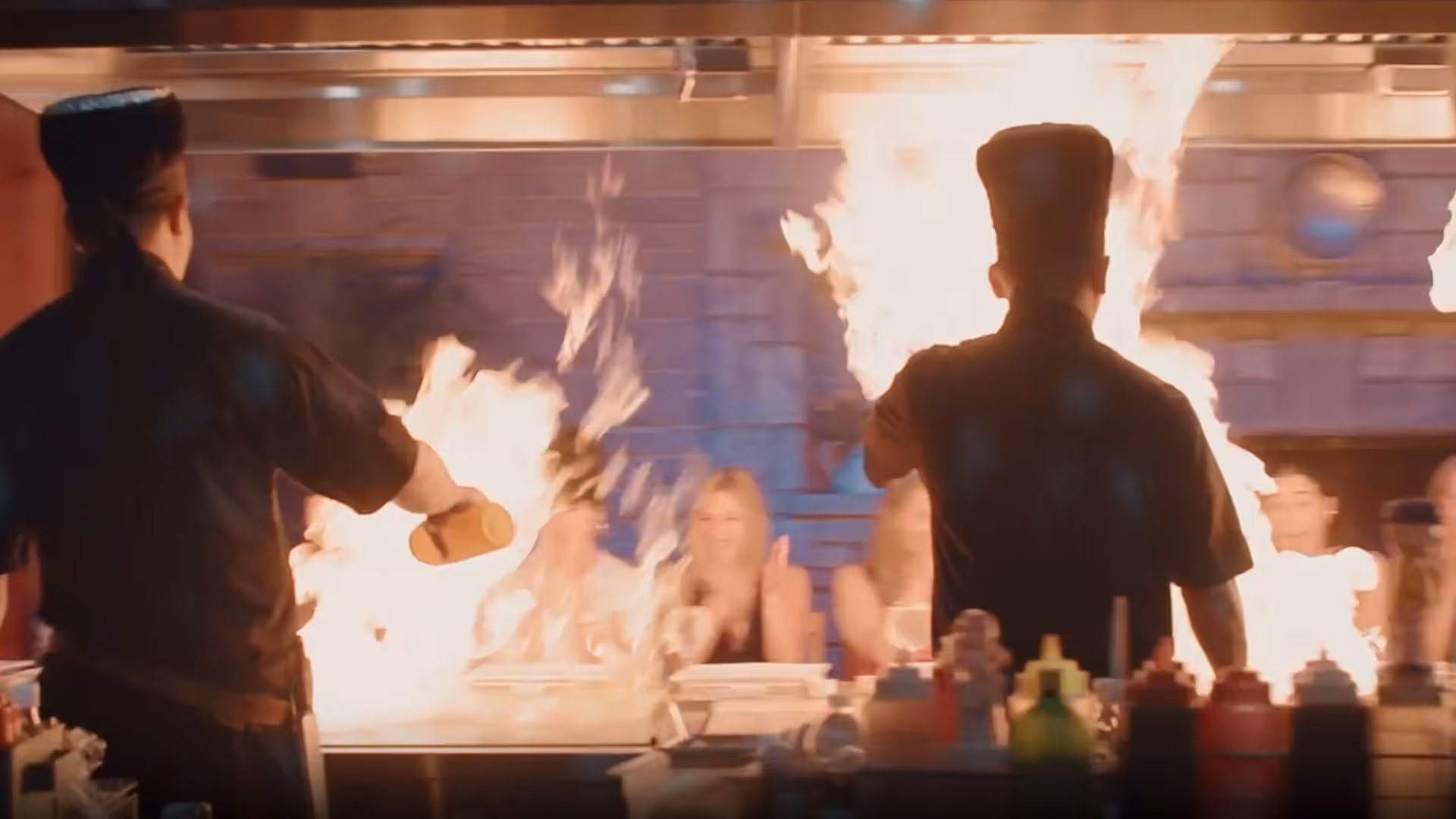 Two chefs in black uniforms performing a teppanyaki cooking show with large flames rising from the grill in front of a seated audience.