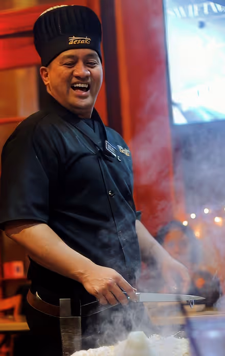 Smiling chef wearing a black uniform and tall black hat with 'desaku' logo, cooking with a spatula in a smoky kitchen environment.