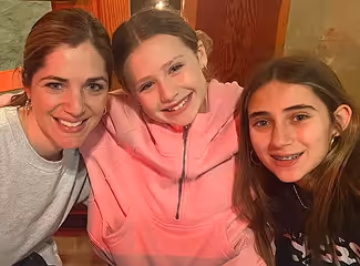 Three smiling females, one adult and two girls, sitting closely together indoors with warm lighting and wooden decor behind them.