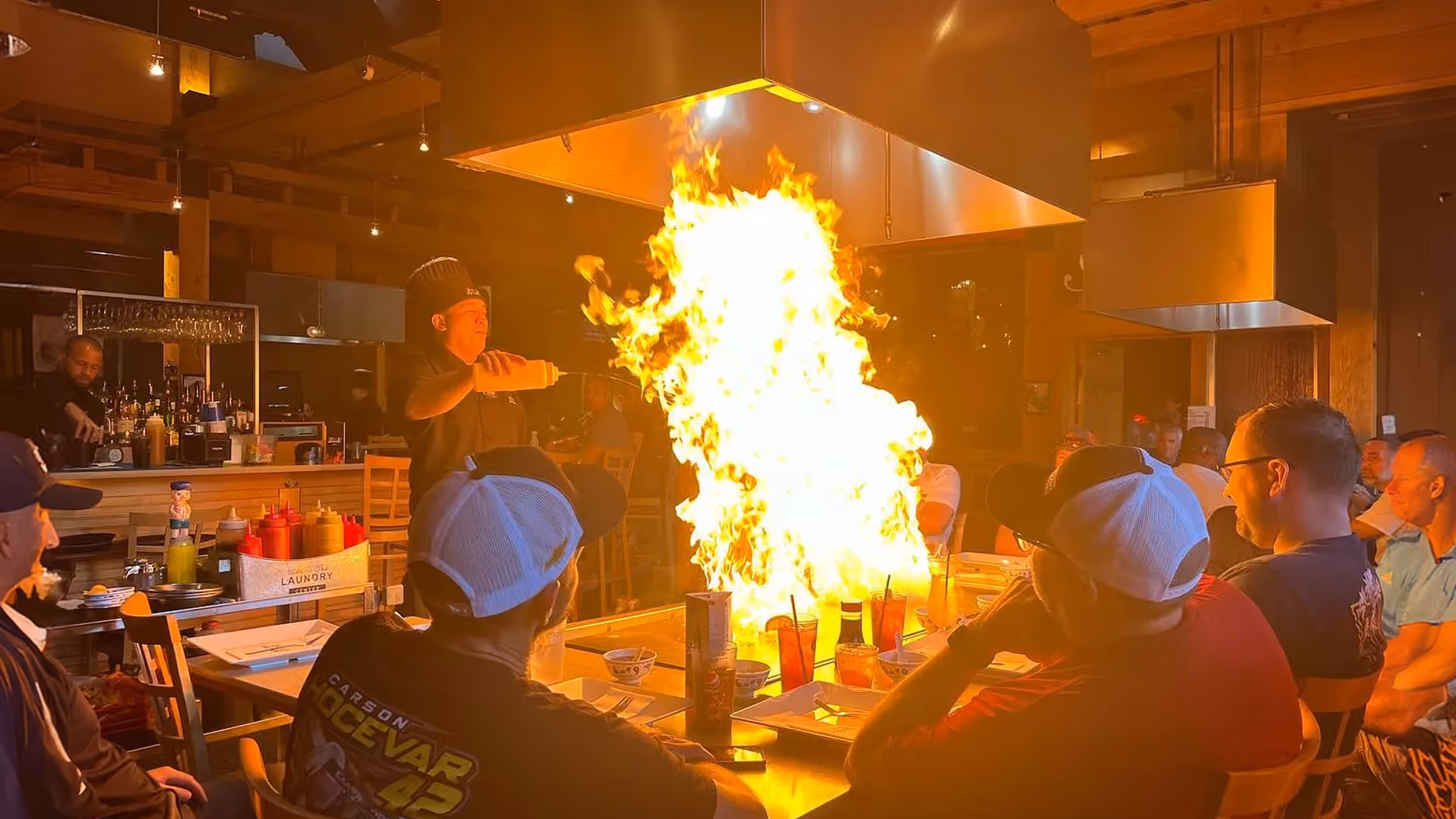 A chef performing a teppanyaki cooking show inside a restaurant, creating a large flame on the grill surrounded by seated diners watching the spectacle.