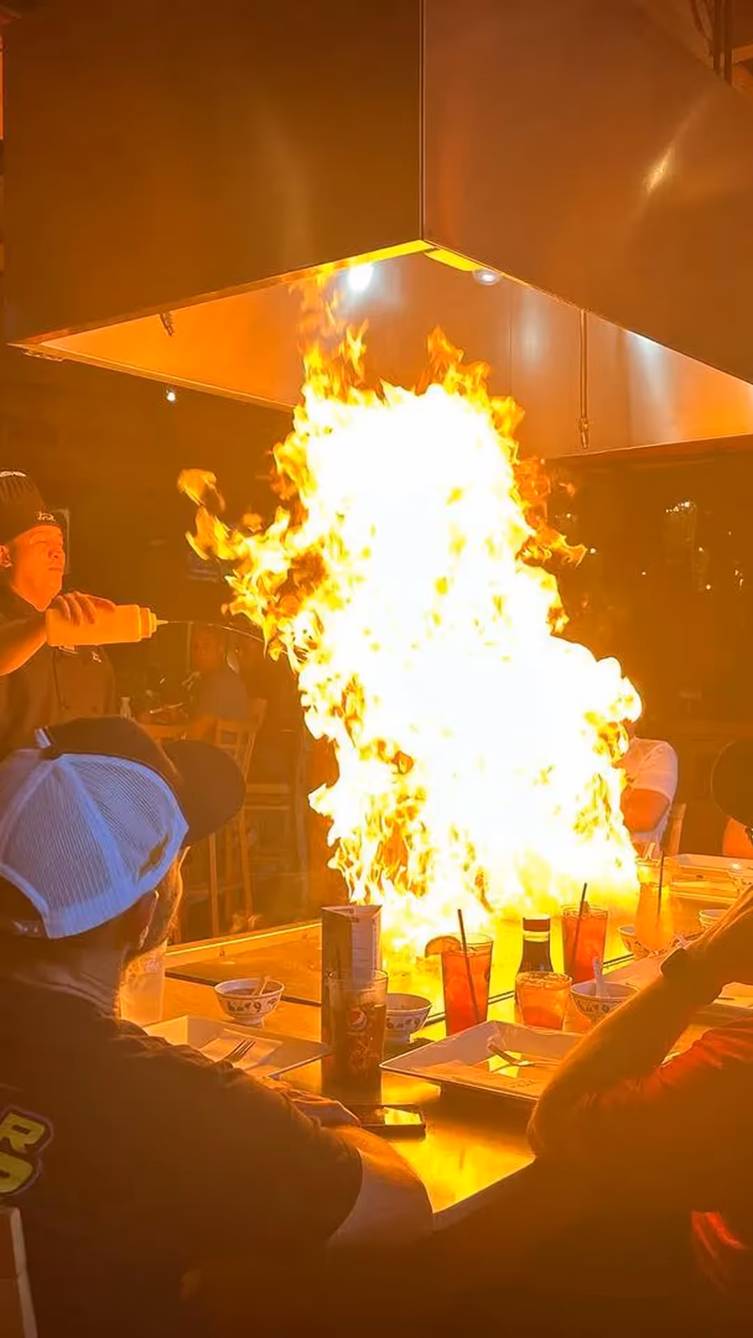 A chef performing a teppanyaki cooking show inside a restaurant, creating a large flame on the grill surrounded by seated diners watching the spectacle.