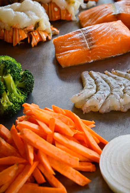 Fresh raw seafood and vegetables arranged on a flat surface, including lobster tails, salmon fillets, shrimp, broccoli florets, sliced carrots, and sliced white onion.