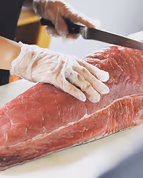 Hands wearing transparent gloves holding and cutting a large raw piece of tuna with a knife on a white surface.