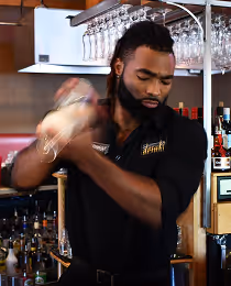 Bartender shaking a cocktail shaker behind a bar with glasses hanging overhead and bottles visible in the background.