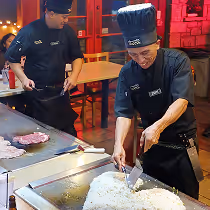 Two chefs in black uniforms cooking on a large flat grill, one preparing rice with a spatula while the other stands nearby with raw meat and cooked steaks on the grill.