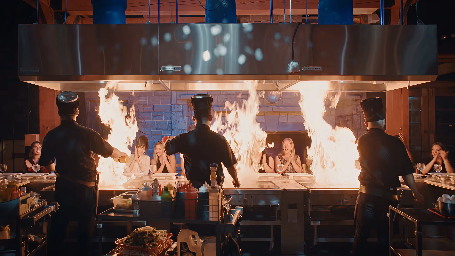 Chef wearing a tall black hat cooking with large flames on a teppanyaki grill while three women seated at the counter watch and clap.