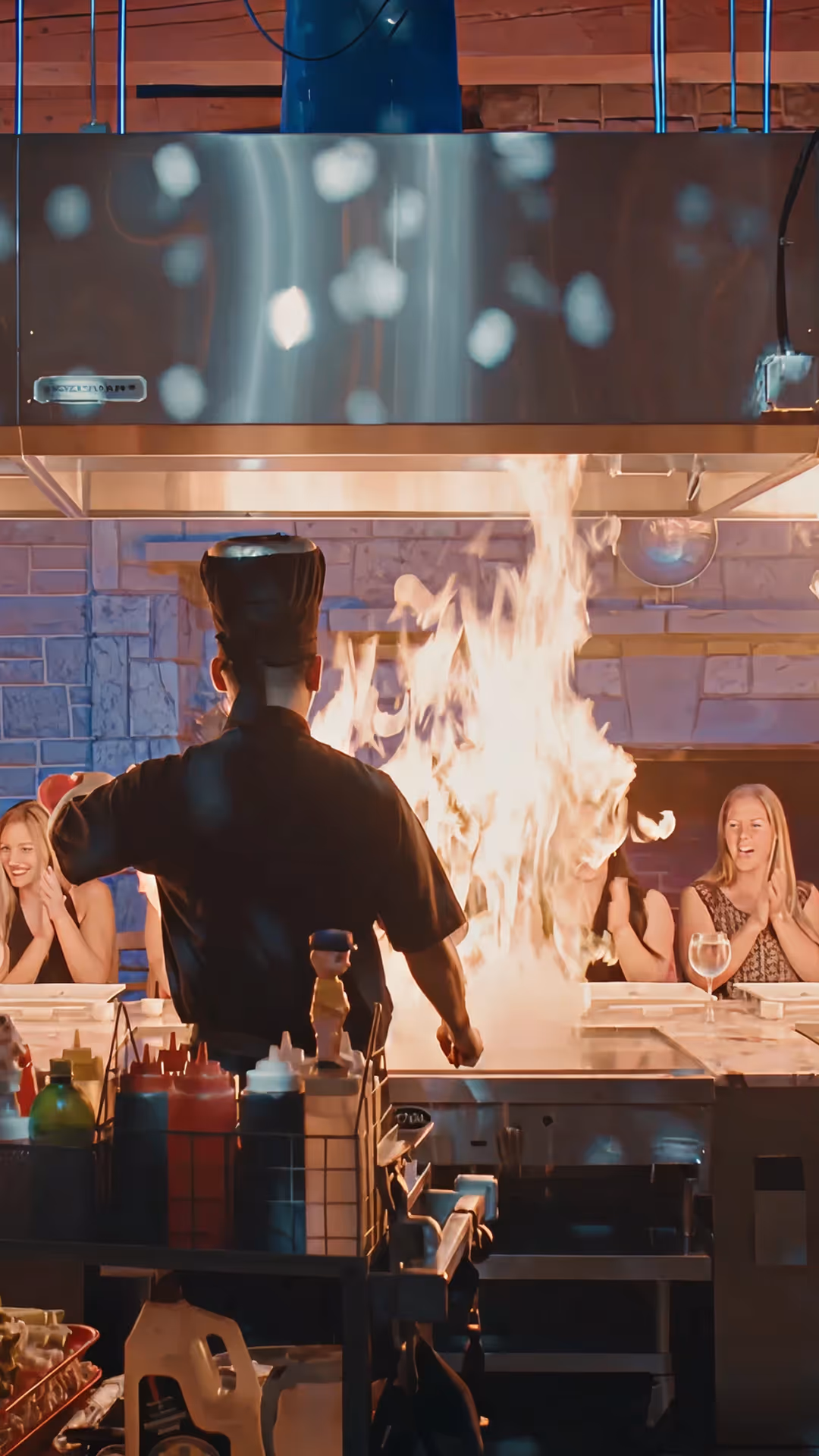 Chef wearing a tall black hat cooking with large flames on a teppanyaki grill while three women seated at the counter watch and clap.