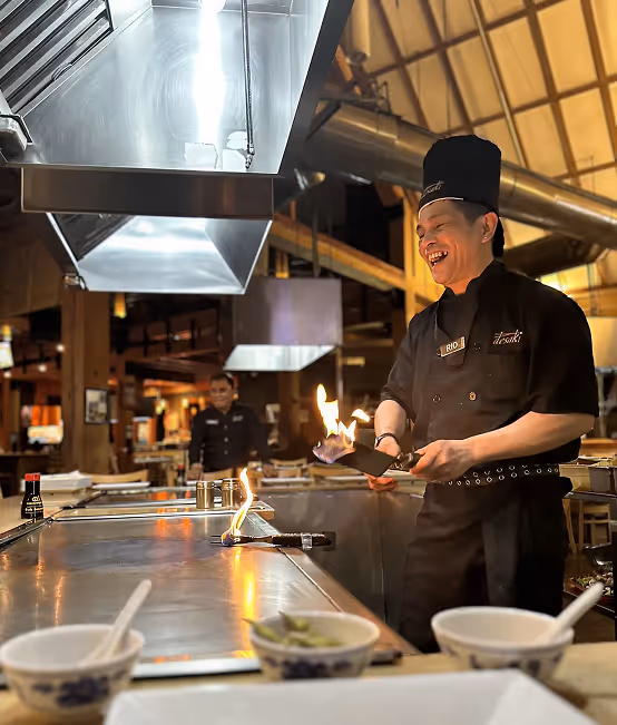 Chef wearing a tall black hat cooking with large flames on a teppanyaki grill while three women seated at the counter watch and clap.