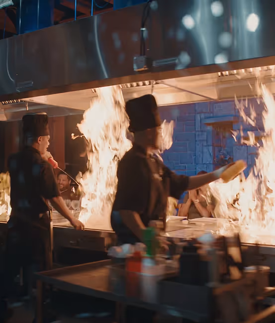 Chef wearing a tall black hat cooking with large flames on a teppanyaki grill while three women seated at the counter watch and clap.