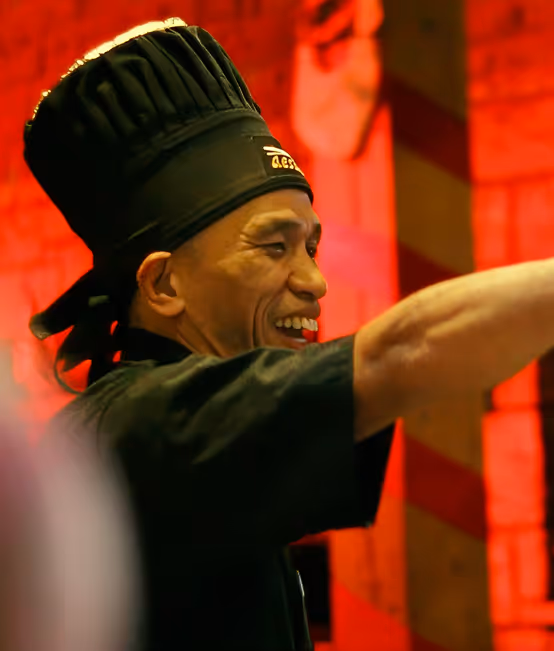 Smiling chef in black uniform and tall black chef hat pointing or gesturing to the right with a warm red-lit background.