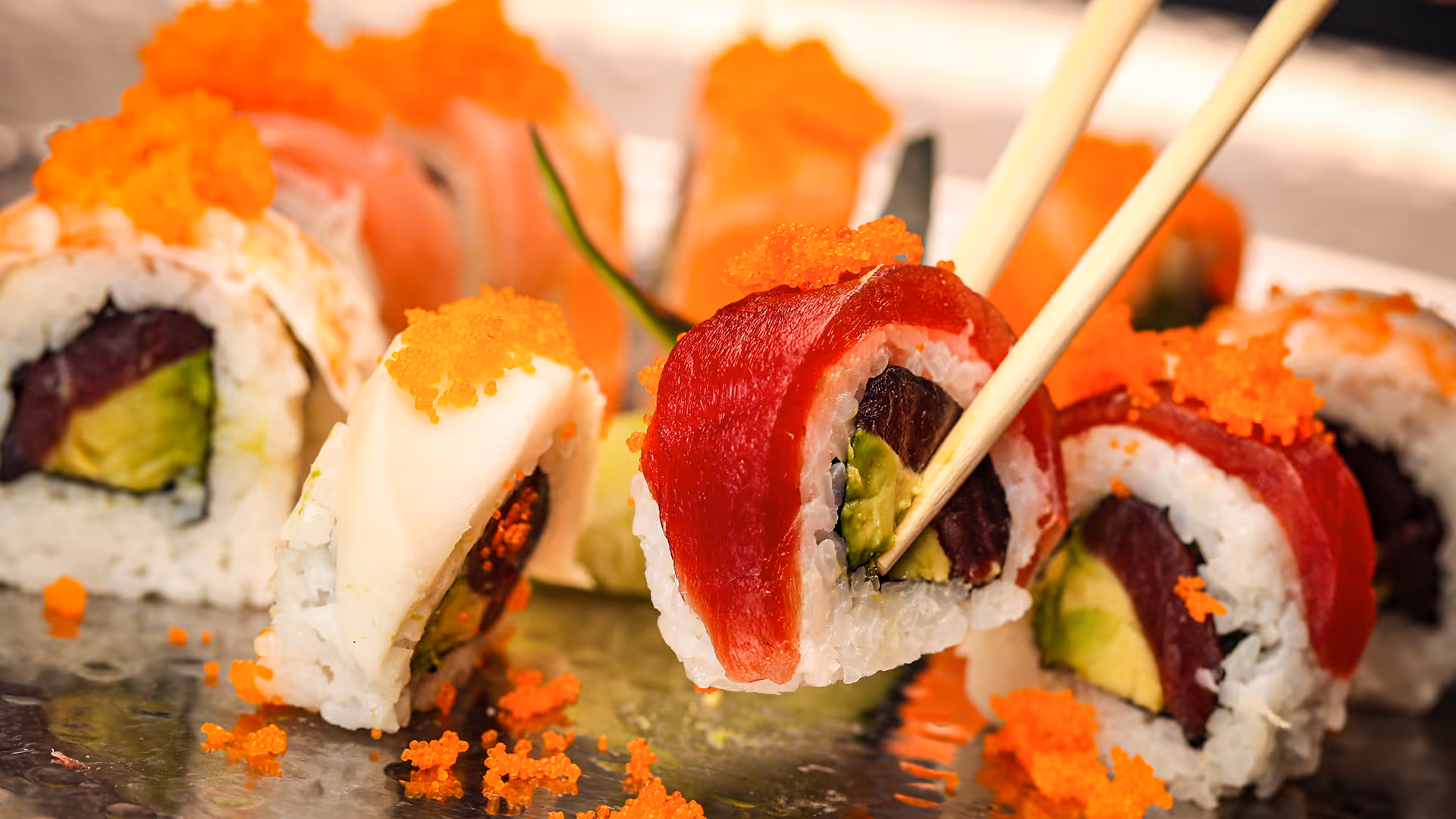 Close-up of chopsticks holding a sushi roll with red tuna, white rice, avocado, and a dark filling, topped with bright orange fish roe, with more sushi and roe blurred in the background.