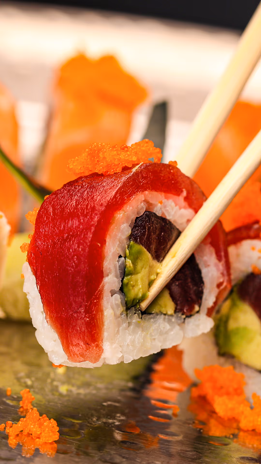 Close-up of chopsticks holding a sushi roll with red tuna, white rice, avocado, and a dark filling, topped with bright orange fish roe, with more sushi and roe blurred in the background.