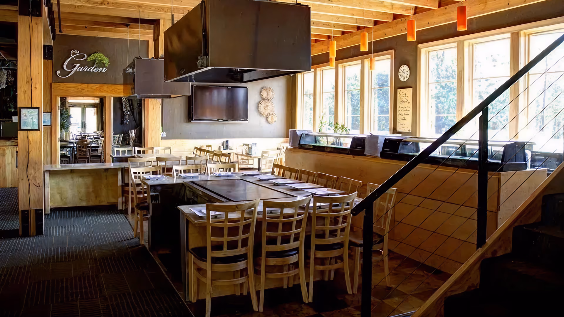 Bright restaurant interior with wooden tables and chairs arranged around a large square grill station under black exhaust hoods, large windows on one side, and a staircase with black railing leading up.
