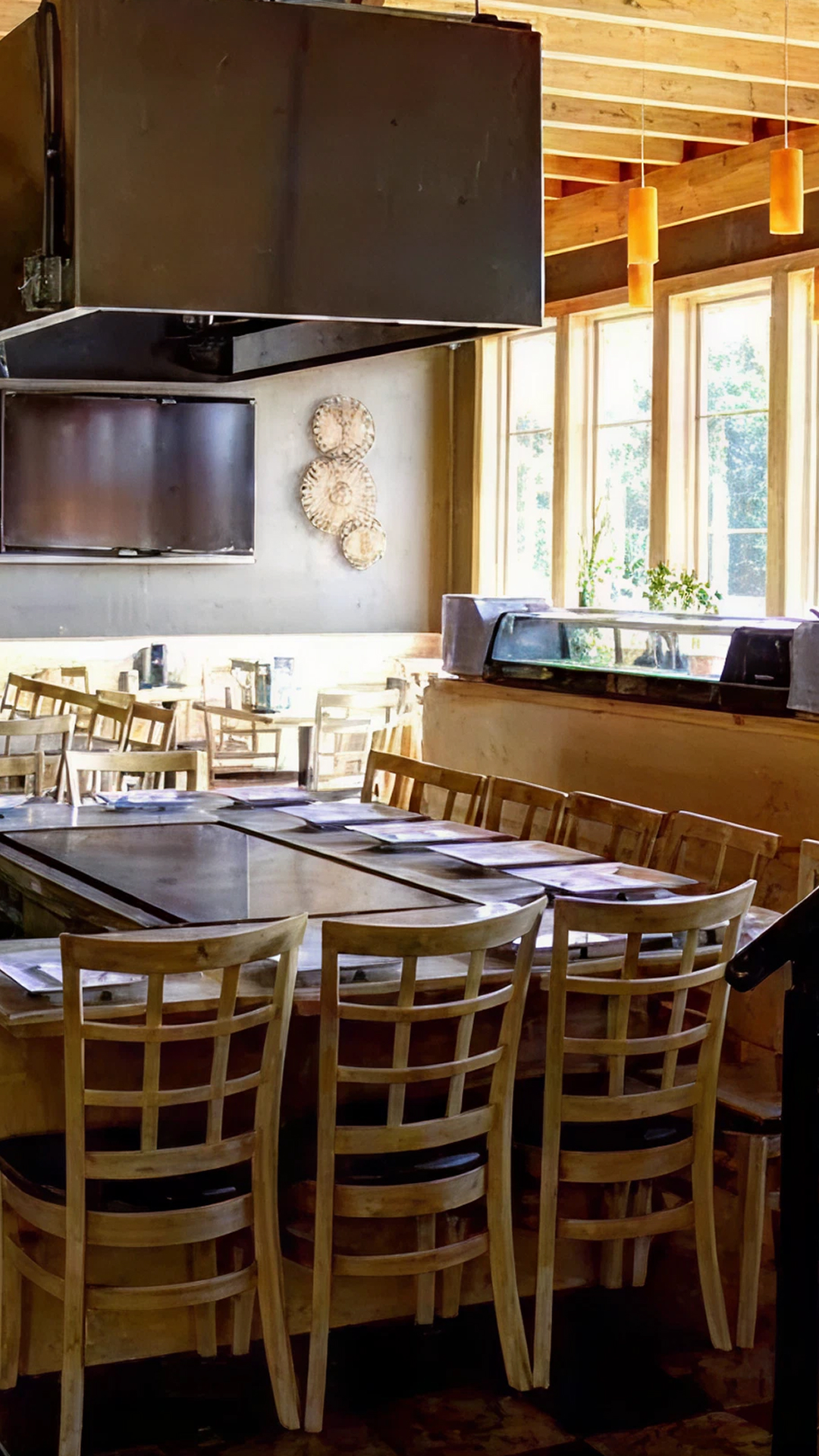 Bright restaurant interior with wooden tables and chairs arranged around a large square grill station under black exhaust hoods, large windows on one side, and a staircase with black railing leading up.