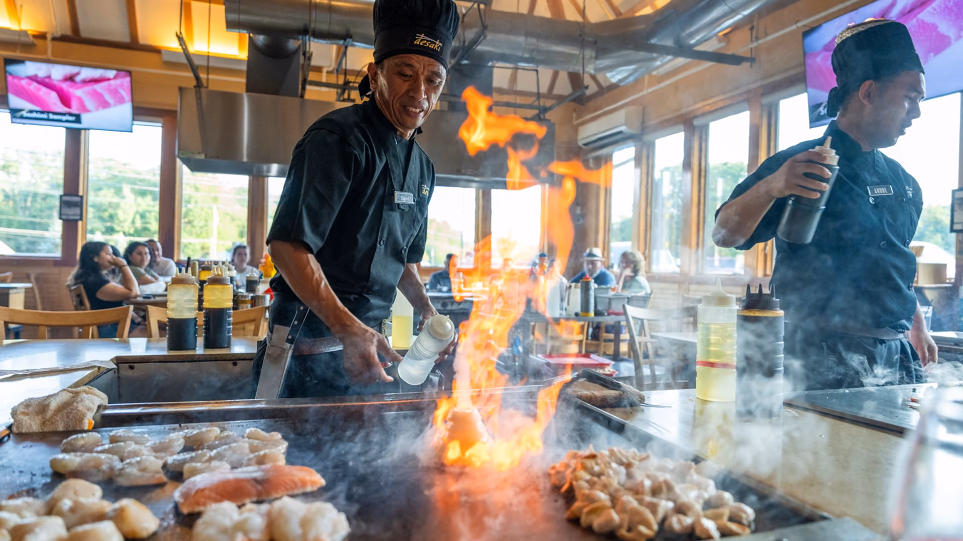 Chef in a black uniform and hat cooking on a hibachi grill with a large flame in a restaurant, with diners visible in the background.