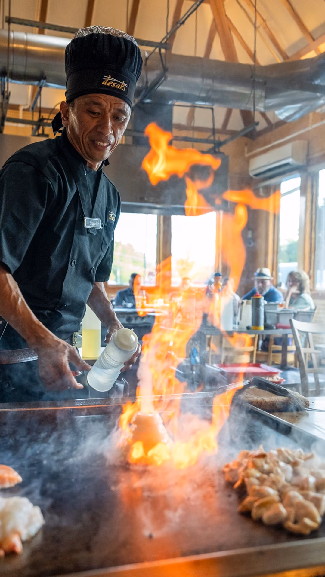 Chef in a black uniform and hat cooking on a hibachi grill with a large flame in a restaurant, with diners visible in the background.