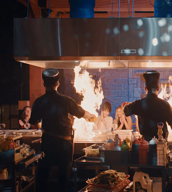 Two chefs performing a teppanyaki cooking show with large flames in front of a group of smiling and clapping diners seated at the counter.
