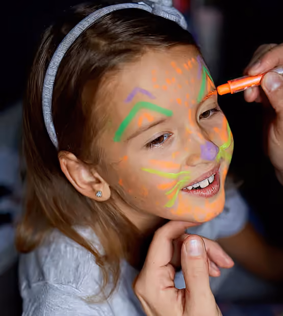 Young girl with a headband having her face painted with bright neon colors, resembling a cat with whiskers and markings, while an adult hand applies orange paint near her eye.