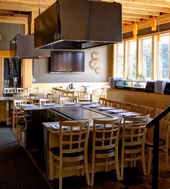 Interior of a restaurant with wooden tables and chairs arranged around a central grill, large windows letting in natural light, and a wall-mounted TV above decorative circular wall hangings.