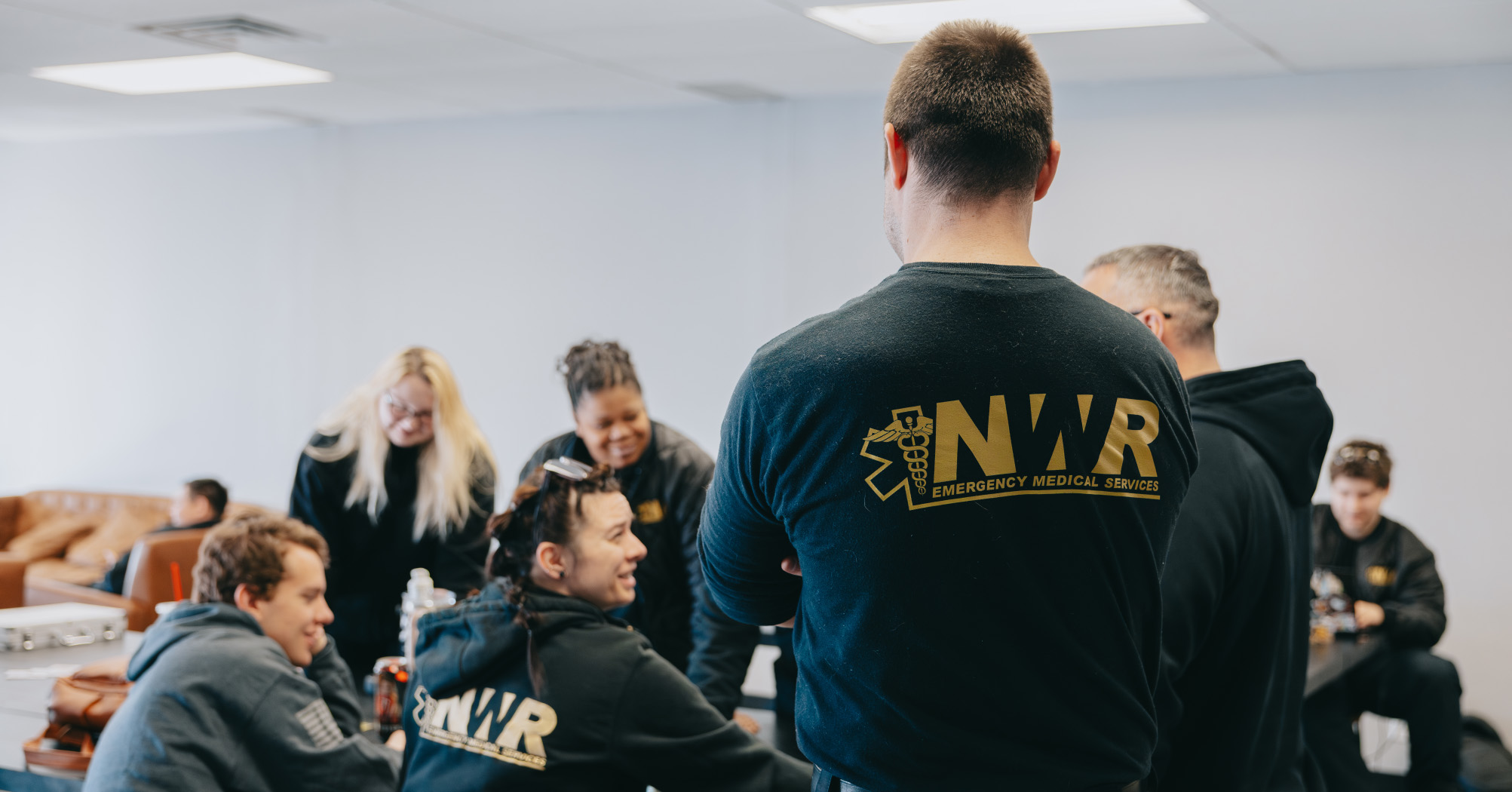 Group of people in a casual meeting room wearing black NWR Emergency Medical Services hoodies, chatting and smiling.