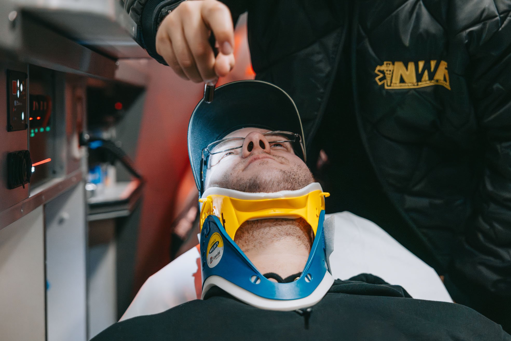 Man wearing glasses and a blue cap with a yellow and blue cervical collar lying down while another person adjusts his head.