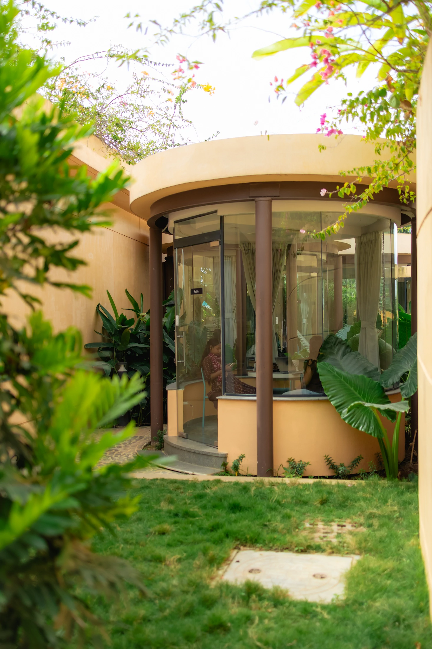 Glass-walled round room with a person seated inside, surrounded by green plants and grass outdoors.