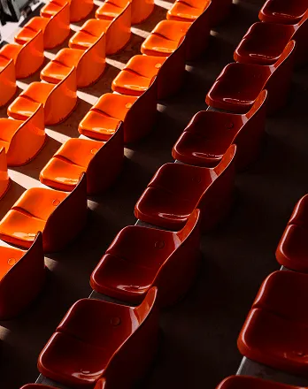 Rows of empty orange stadium seats lit by sunlight with shadows in between.