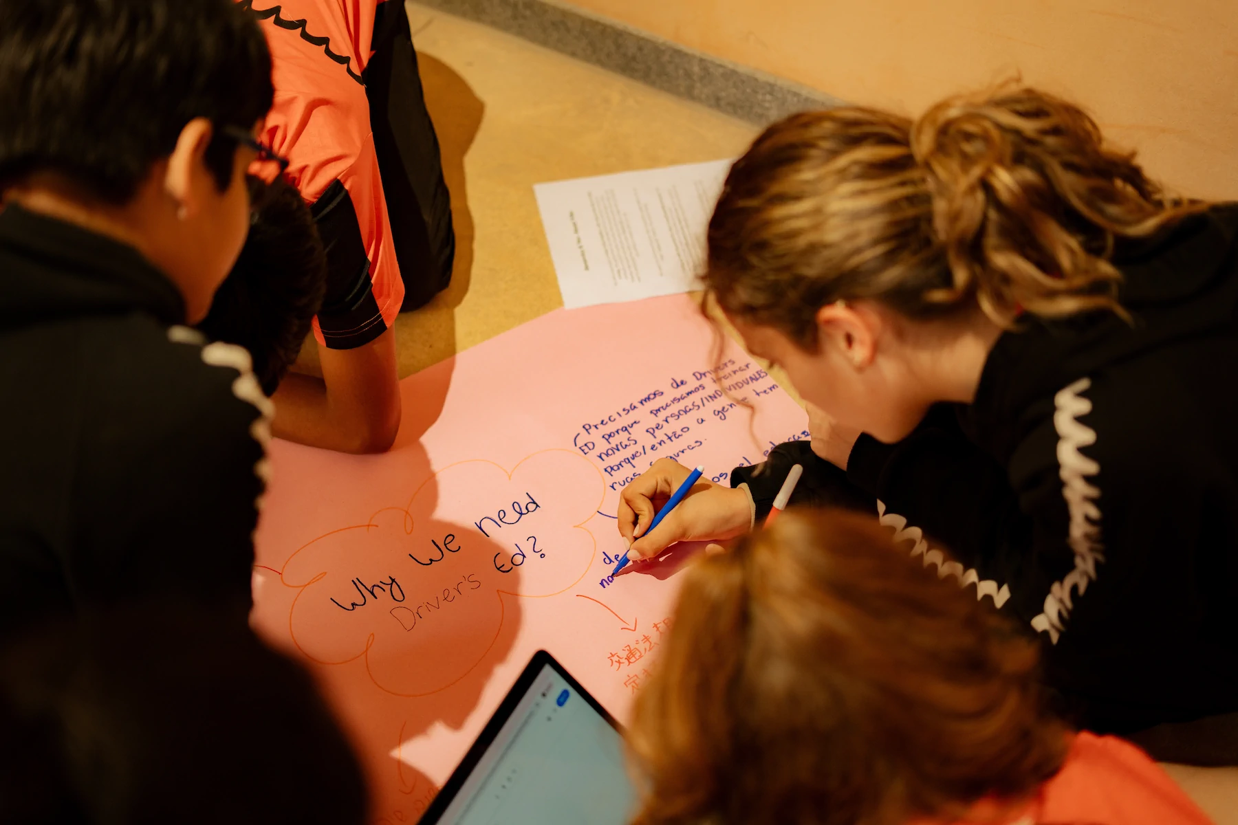 Several young people write reasons on a large paper titled 'Why we need Driver's Ed?'