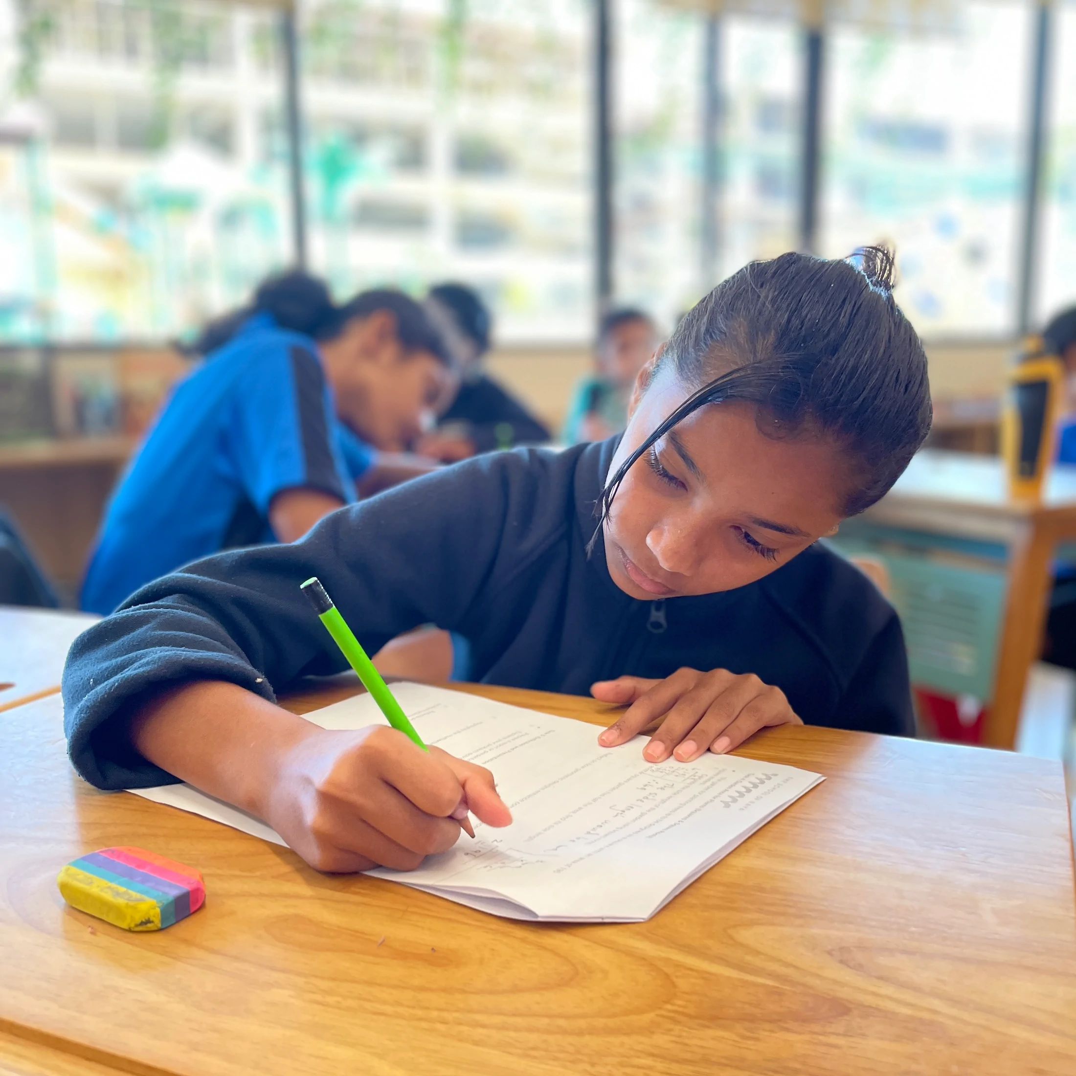 Student in a classroom focused on writing on a worksheet with a green pencil.