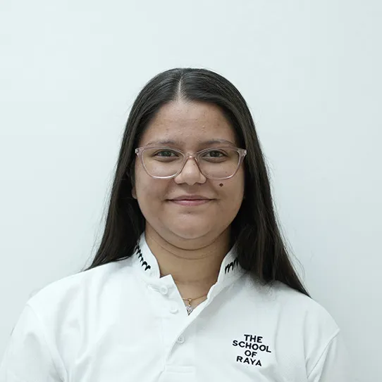 Woman with long dark hair and glasses wearing a white shirt with The School of Raya logo.
