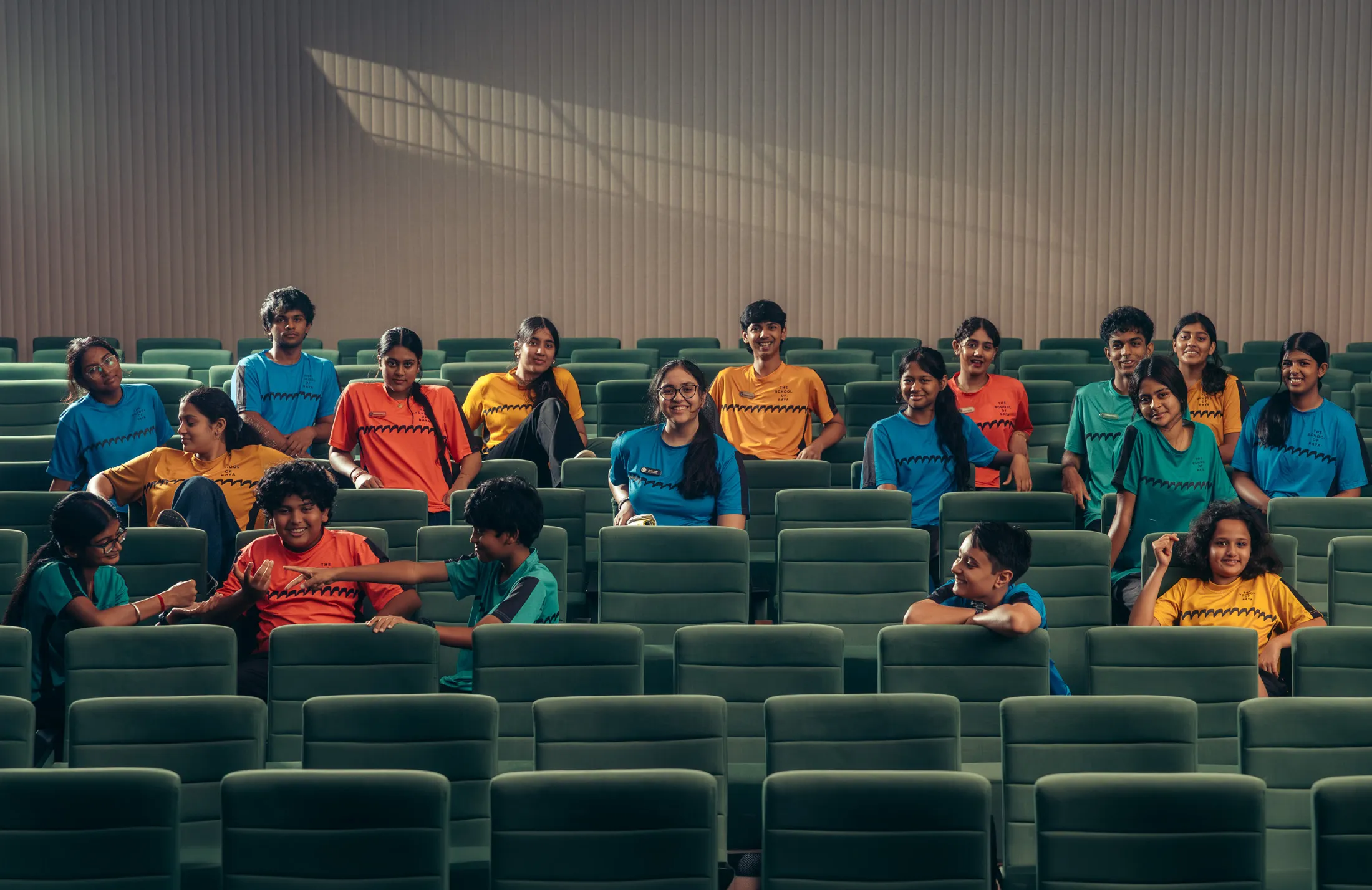 Group of teenagers in colorful shirts sitting in green auditorium seats, some interacting and smiling.
