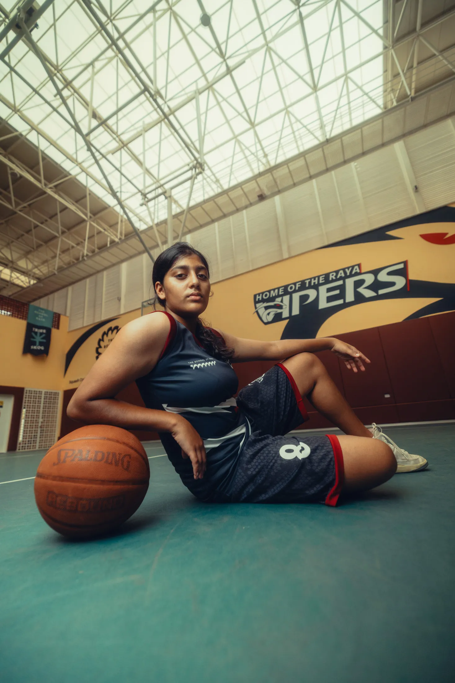 Female basketball player in jersey sitting on court floor with ball inside an indoor gym.