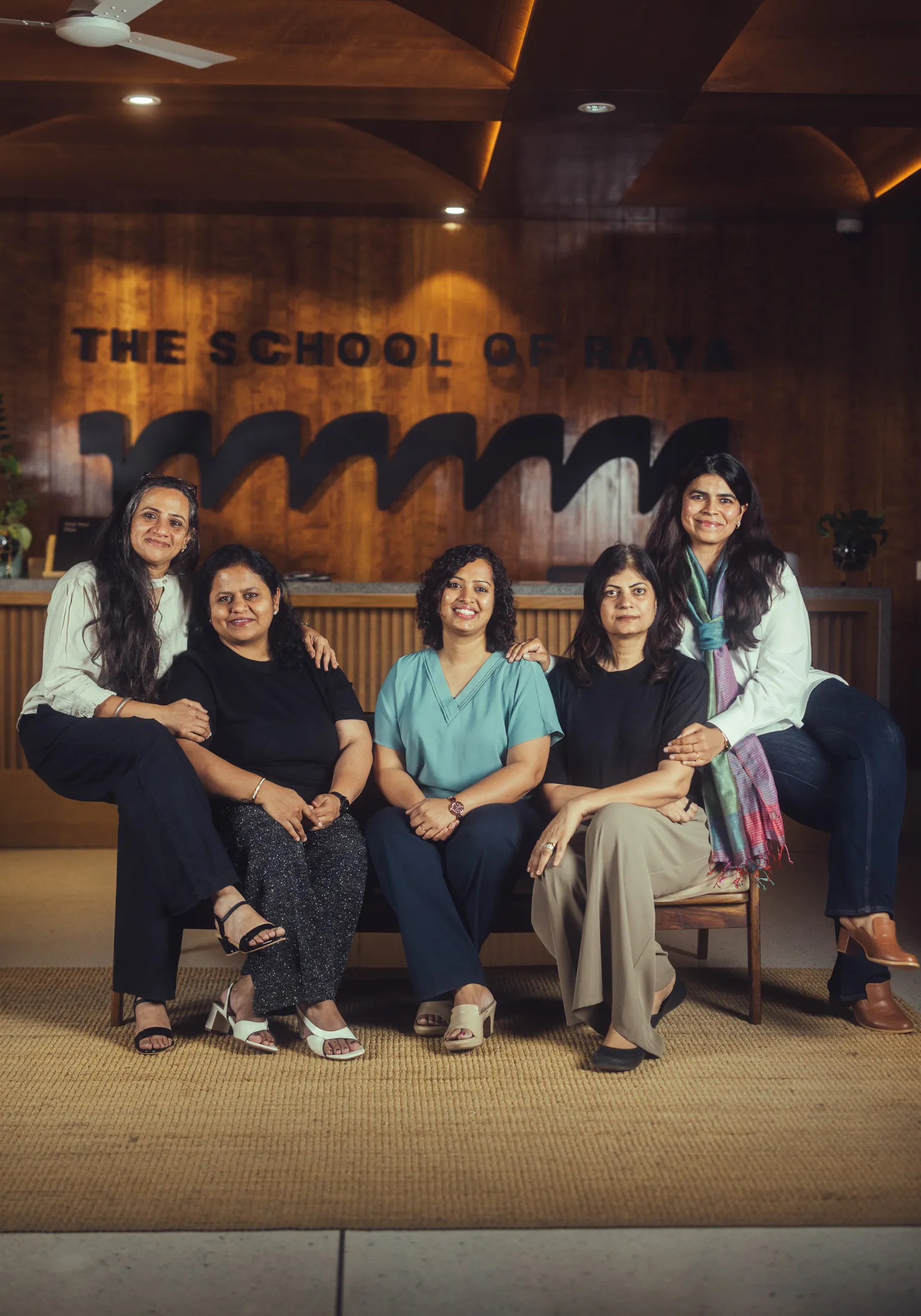 Five women sitting and smiling in front of a wooden wall that says The School of Raya.