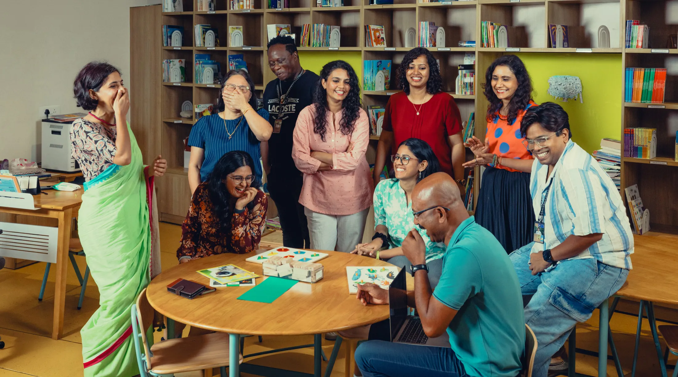 Group of adults smiling and laughing around a table in a room with bookshelves and educational materials.