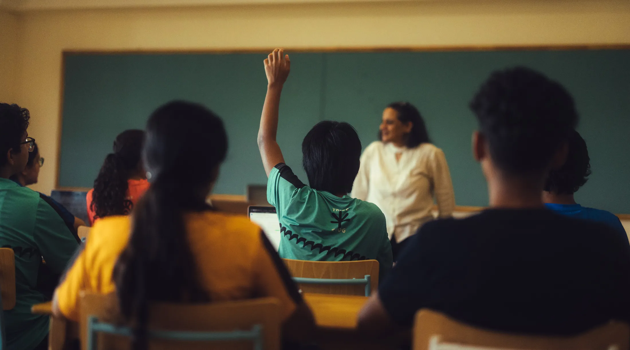 Student raising hand in classroom with teacher standing near chalkboard and other students seated.