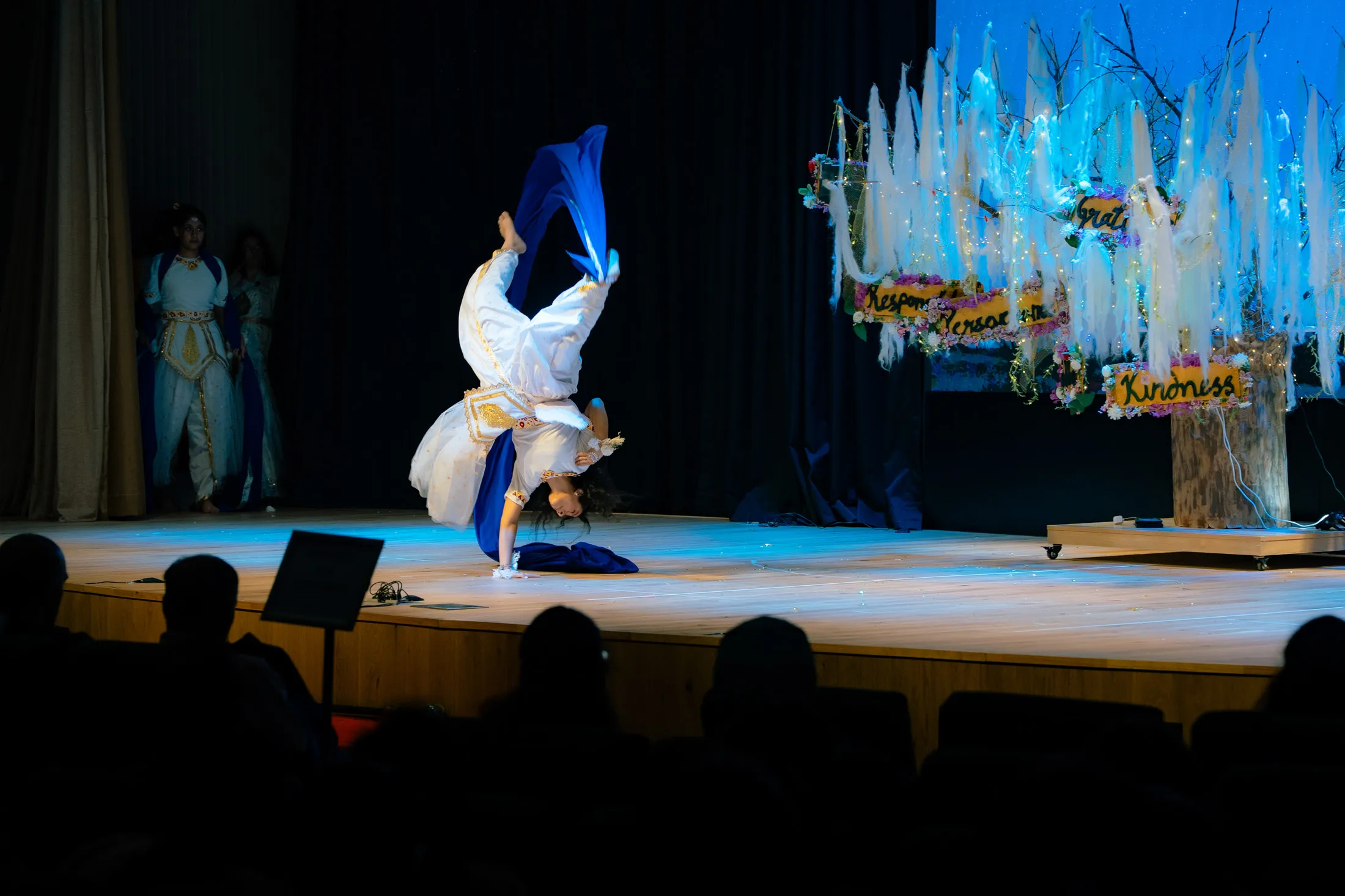 Performer in white and gold costume doing a handstand on stage during a theatrical dance show.