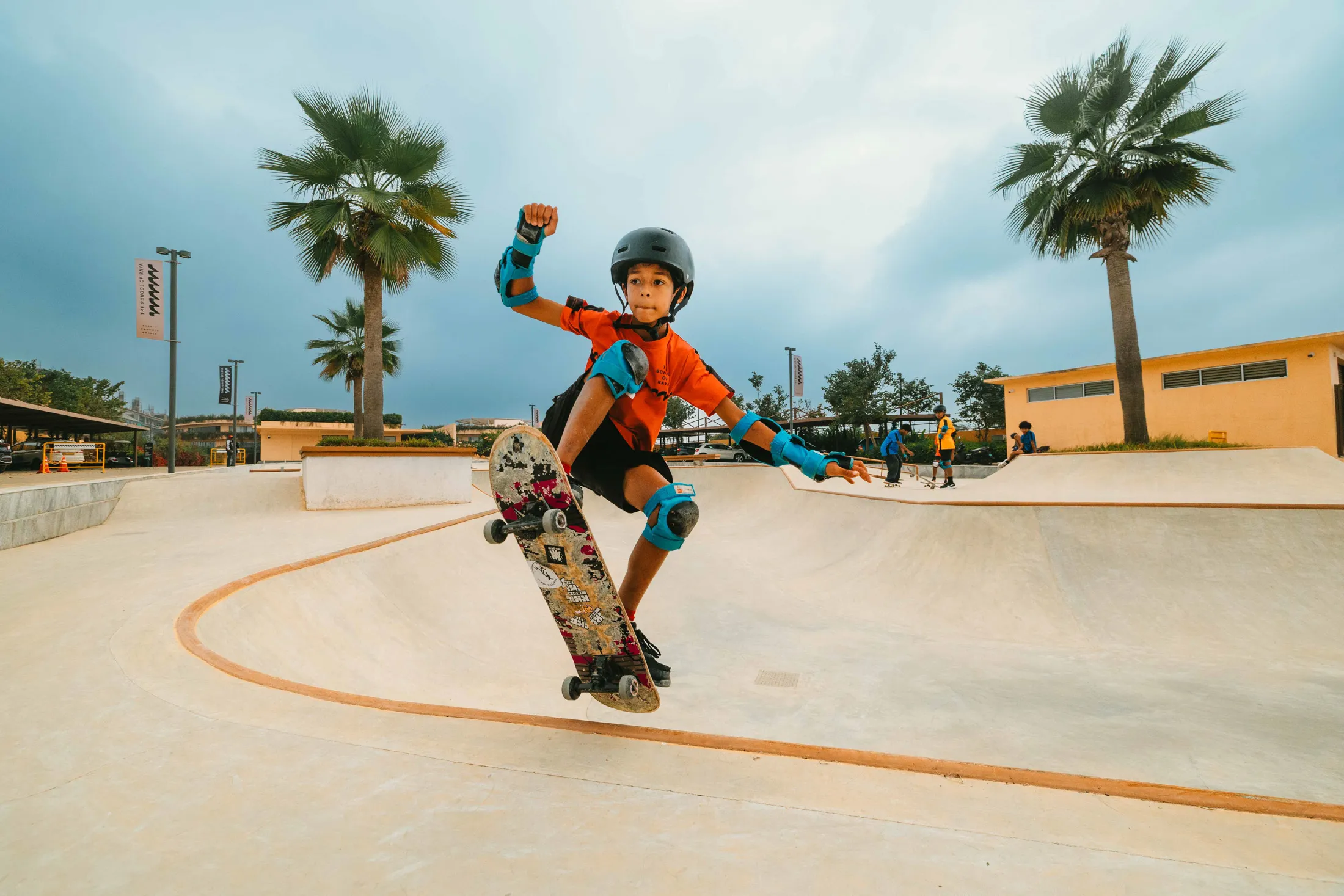 Young boy in helmet and pads performing a skateboard trick at a skate park with palm trees and cloudy sky.