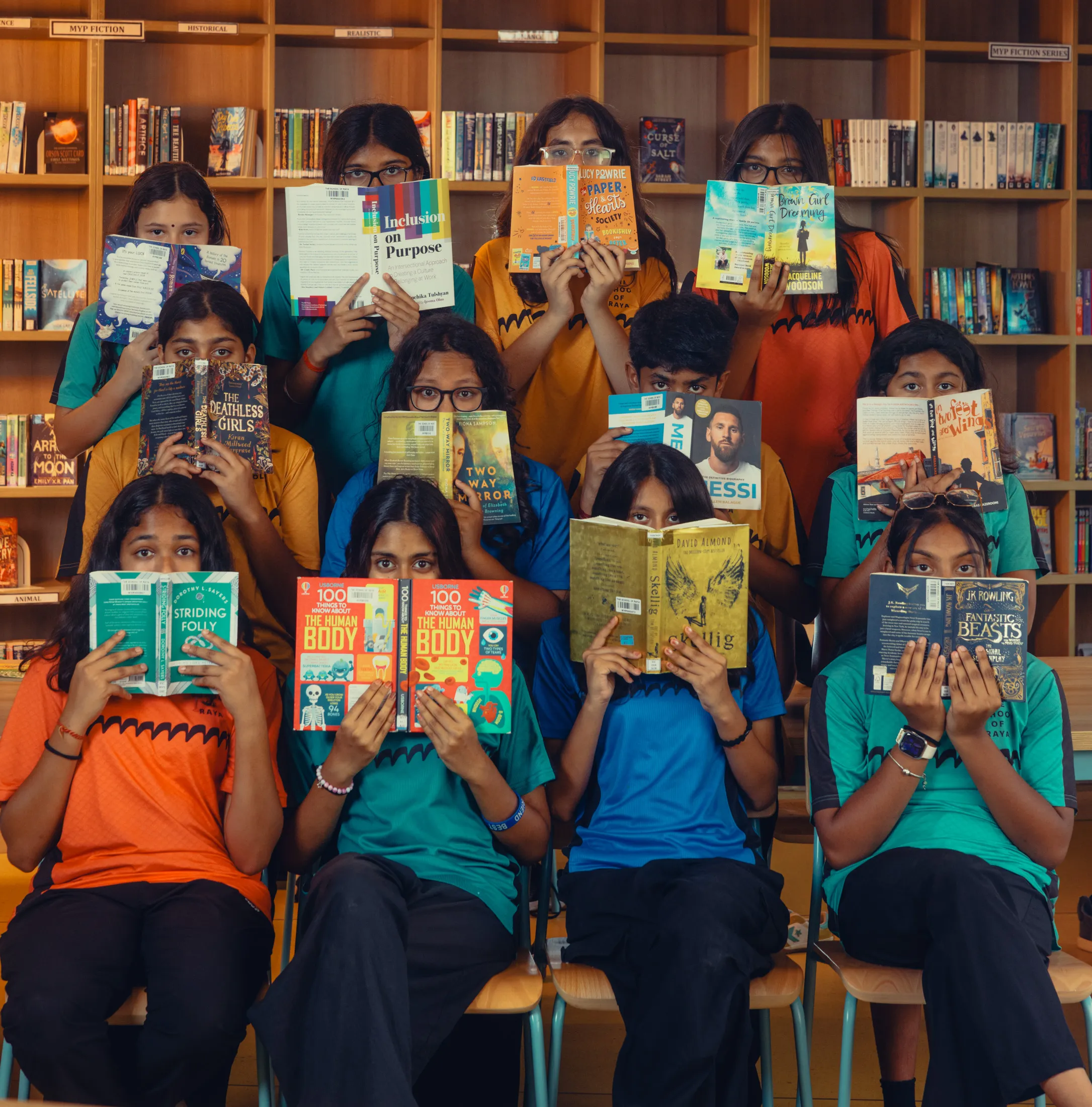A group of students sitting and standing in a library holding books in front of their faces.
