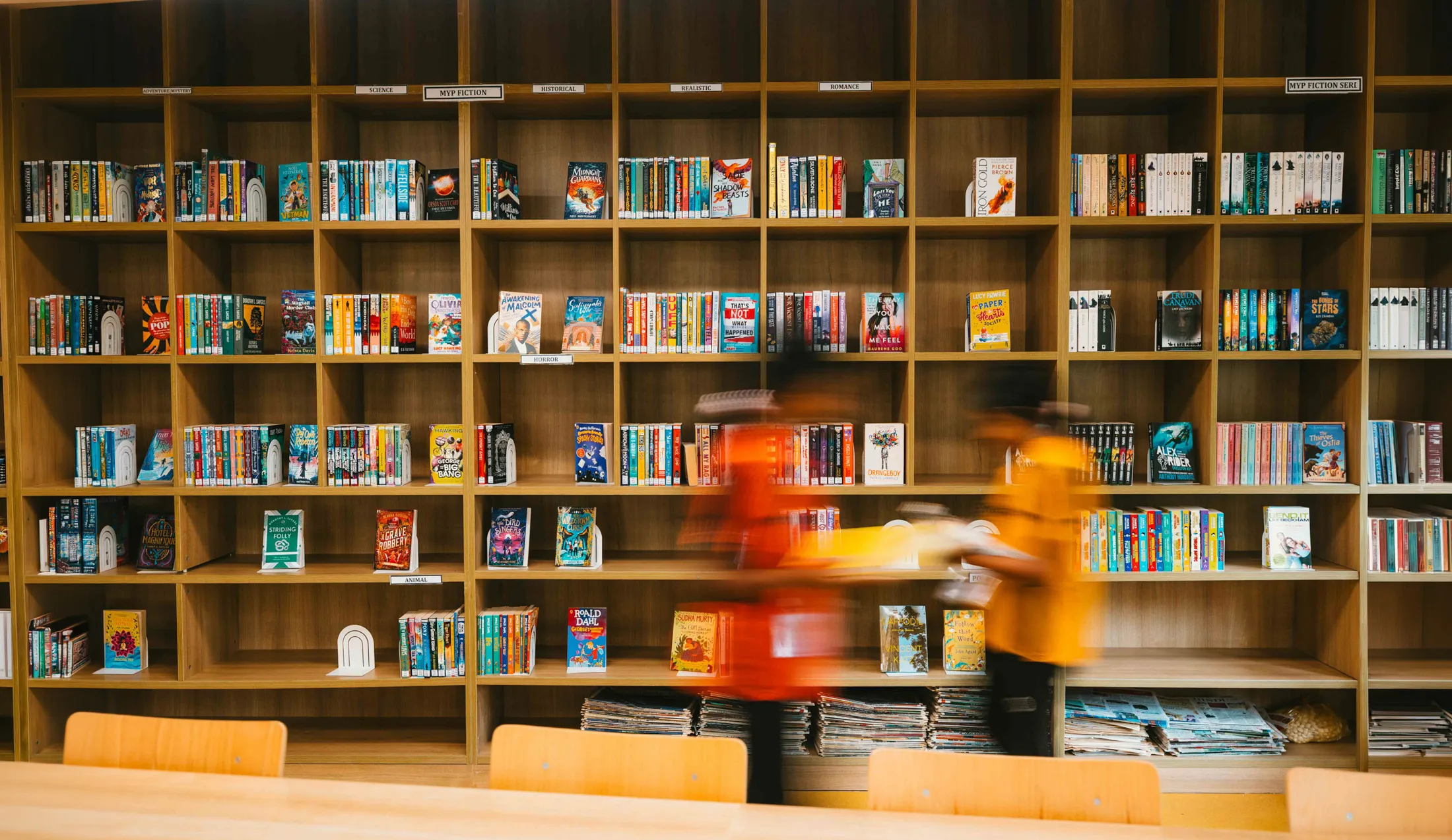 Bookshelf filled with books labeled by genres and two blurred people passing in front in a library.