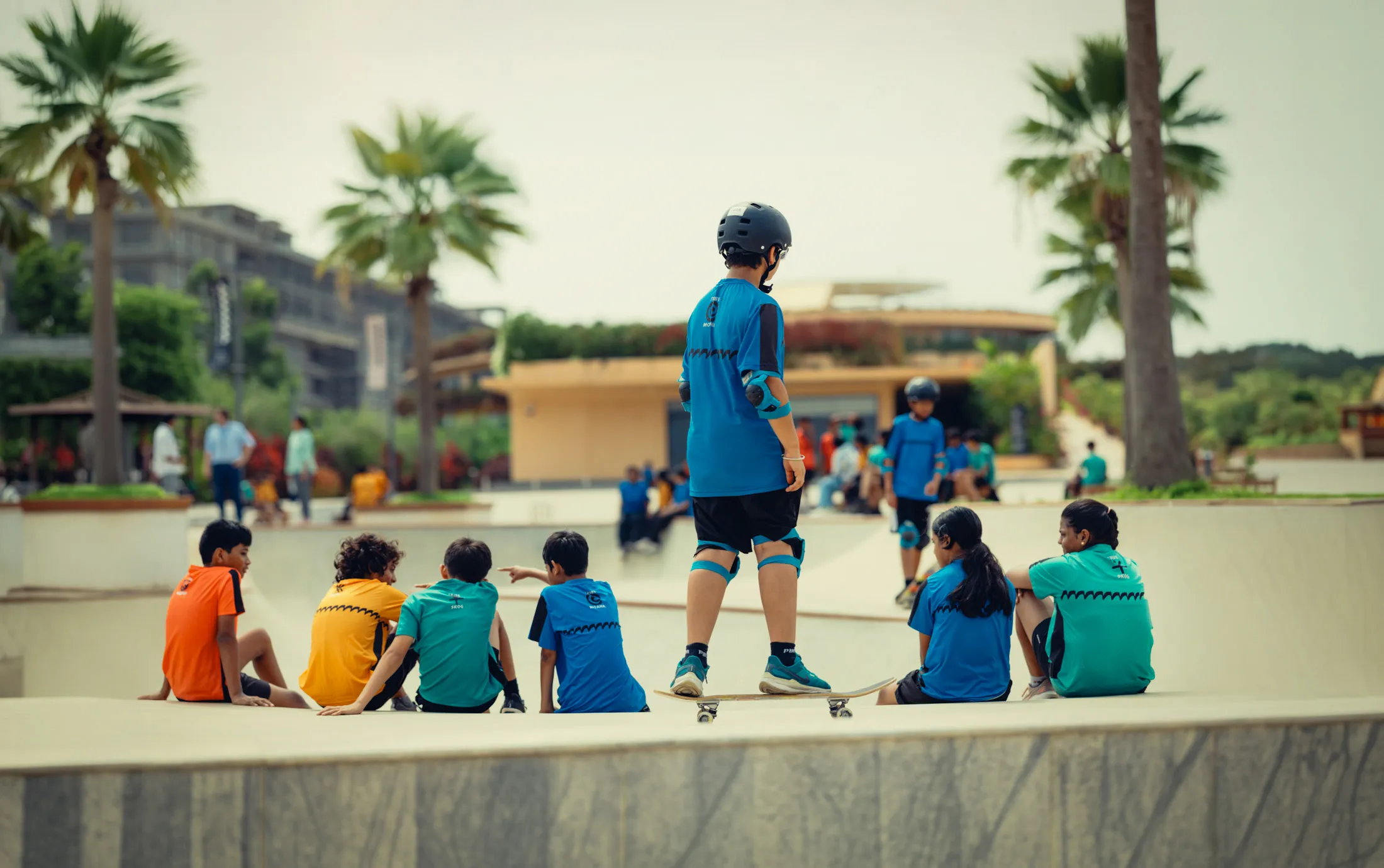 Group of children in colorful shirts at a skate park, one boy on skateboard wearing helmet and pads.