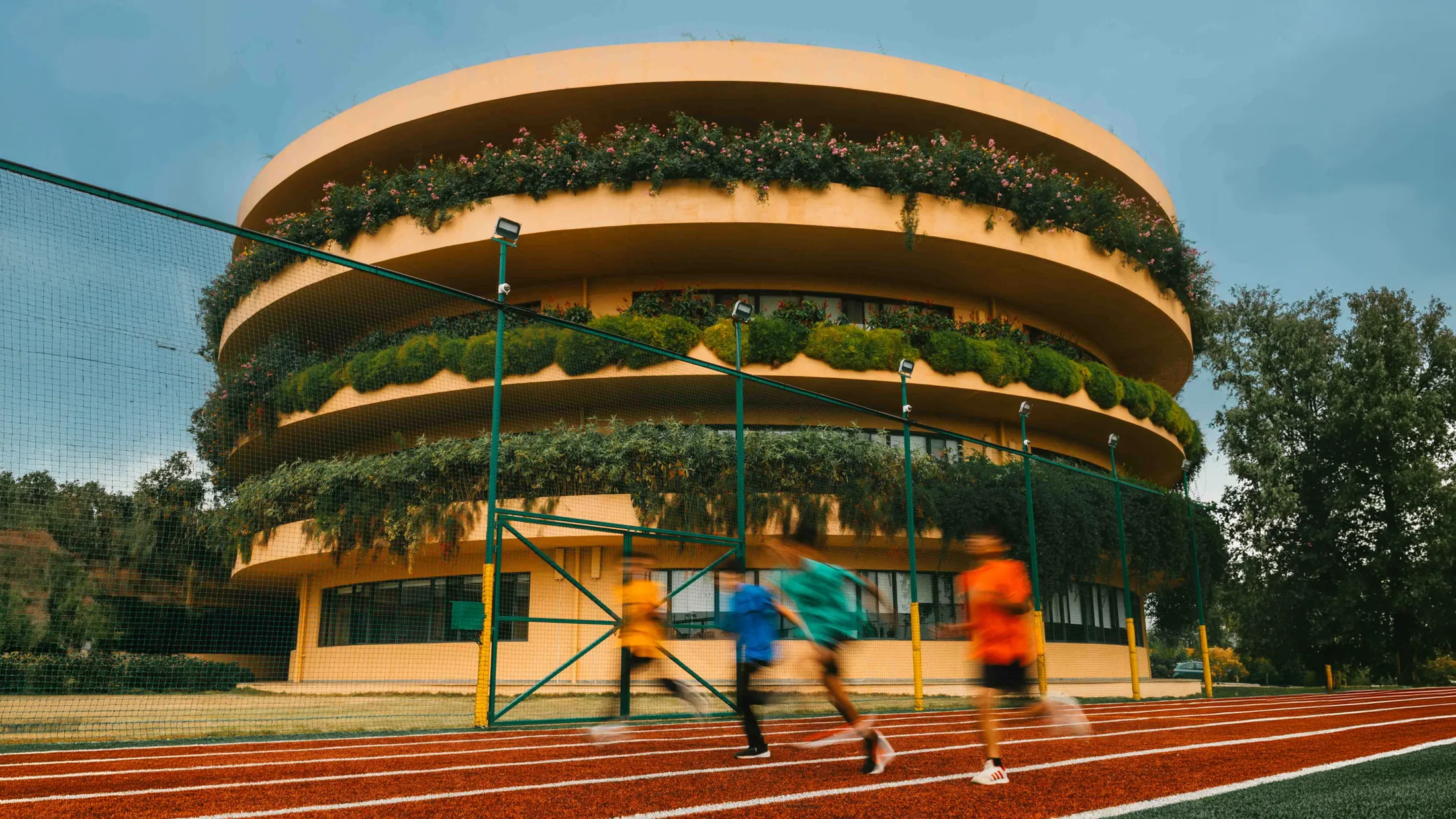 Blurred children running on red track in front of a round building with green plants on balconies.