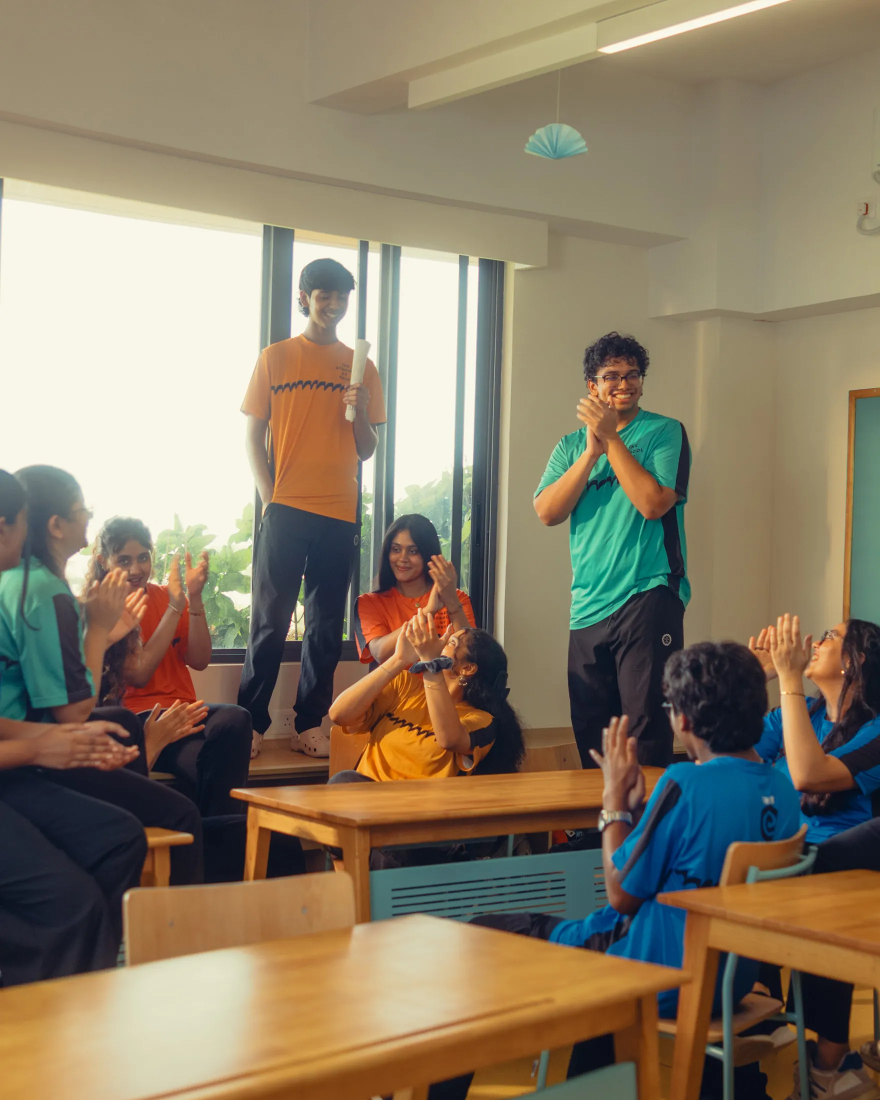 Group of students in colorful shirts clapping and smiling in a classroom with wooden desks.