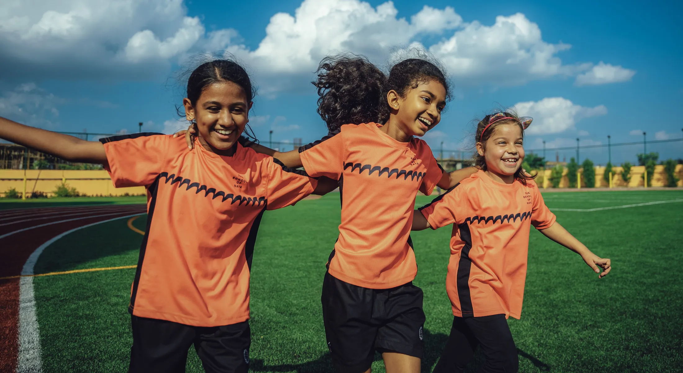 Three girls in orange sports jerseys smiling and walking arm-in-arm on a green sports field.