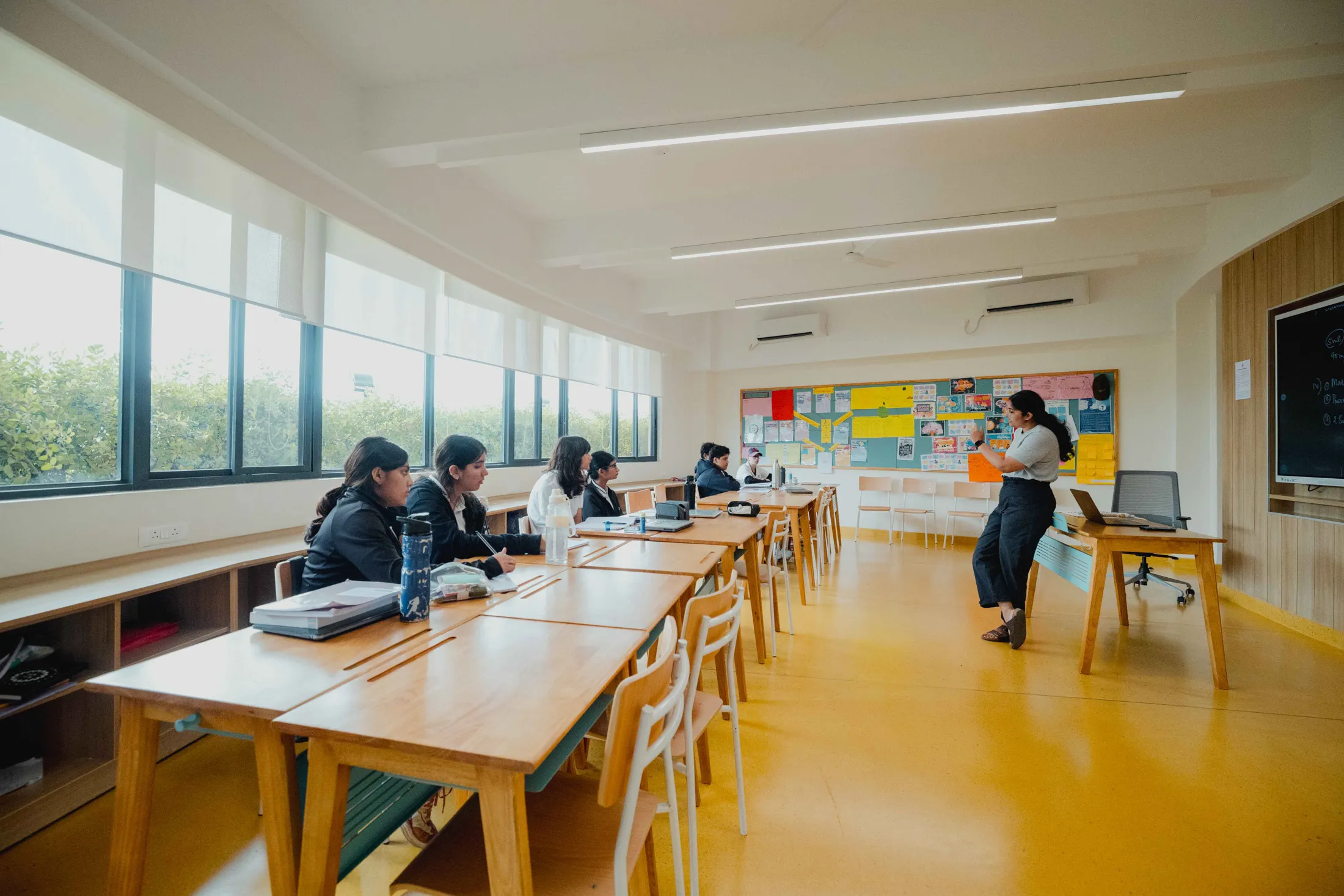 Students seated at wooden desks in a bright classroom while a teacher stands near a desk.