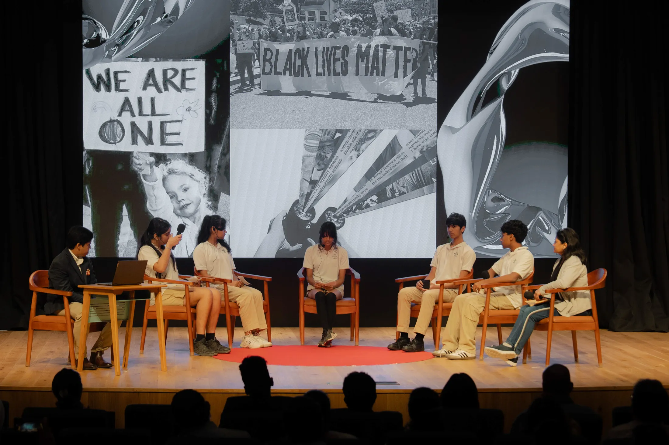 Seven people sit on stage chairs in a discussion with Black Lives Matter images on a large screen behind them.